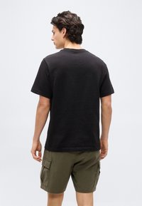 Άνδρας γυρισμένος προς τα πίσω, φορά μια απλή μαύρη κοντομάνικη μπλούζα και χακί cargo σορτς, μπροστά σε απλό ανοιχτό φωτεινό φόντο.