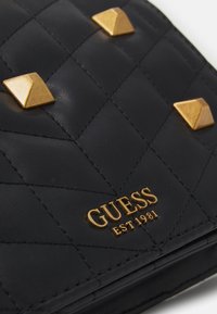 Sort quiltet lædertaske med guldfarvede pyramide-studs og præget brandlogo med "EST 1981" i bunden.