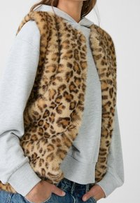Weste aus Kunstfell im Leopardenmuster, die über einem grauen Hoodie getragen wird. Die Weste hat eine weiche Textur, einen runden Ausschnitt und ein offenes Design.