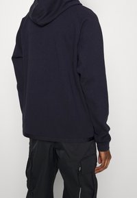 Granatowy hoodie o luźnym kroju, długich rękawach i kapturem ze sznurkiem. Materiał wydaje się gładki, z minimalnymi detalami przeszyć.