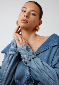 Jasnoniebieska, oversize'owa kurtka dżinsowa z dużym kołnierzem i podwiniętymi rękawami, z guzikiem na mankiecie. Modelka nosi złote kolczyki-koła.