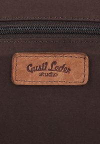 Bruine stof met een zwarte ritssluiting boven een gestikte rechthoekige leren patch met de tekst "Gusti Leder studio."