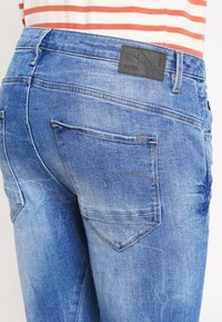 Jeans de mezclilla azul desgastados con un ajuste de media cintura, que presentan dos bolsillos traseros y un parche de piel negra de la marca en la cinturilla.