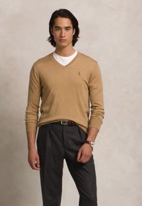 Pull beige en V en tissu léger, avec des manches longues et un petit logo sur la poitrine, associé à un pantalon gris ajusté.