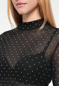 Sort gennemsigtig top med høj hals, der har et polkaprikket mønster af skinnende rhinsten, og en tætsiddende pasform over en sort bh under.