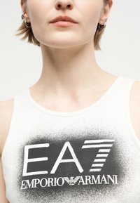 Κοντινό πλάνο ενός ατόμου που φοράει λευκό αμάνικο με το λογότυπο "EA7 Emporio Armani" σε μαύρο στυλ γραφής με σπρέι στο στήθος.
