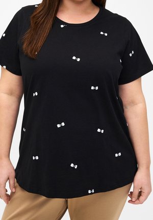 Vrouw draagt een zwart shirt met korte mouwen en kleine witte strikpatronen, gecombineerd met beige broek, tegen een effen achtergrond.