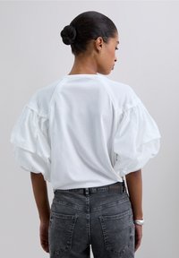Camiseta blanca de algodón con mangas voluminosas y abullonadas, con un cuello redondo y un diseño relajado en la parte posterior, combinada con jeans de mezclilla grises.