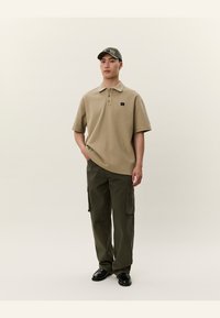 Mann iført en beige polo-skjorte, mørkegrønne cargo-bukser, svarte sko og en grønn baseballcaps som står foran en ensfarget bakgrunn.