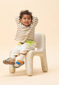 Ein Kind sitzt auf einem gesprenkelten, hellen Stuhl und trägt ein beige-weiß gestreiftes Hemd, texturierte, cremfarbene Hose mit grünen Akzenten und bunte Schuhe.