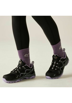 Czarne i fioletowe buty trekkingowe noszone z fioletowymi skarpetkami i czarnymi legginsami, pokazane od dolnej części nóg w dół na jednolitym tle.
