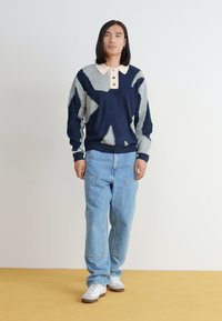 Σκούρο μπλε και ανοιχτό γκρι πλεκτή μπλούζα με μπεζ γιακά, συνδυασμένη με ανοιχτό μπλε τζιν παντελόνι και λευκά αθλητικά παπούτσια πάνω σε κίτρινο πάτωμα.