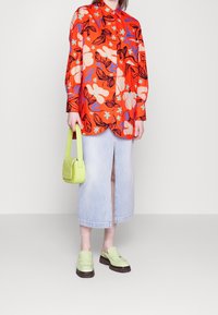Chemise à boutons rouge à fleurs avec une coupe décontractée, associée à une jupe en denim bleu clair. Les accessoires comprennent un petit sac vert citron et des sabots assortis.