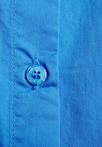 Tecido azul com uma textura suave, apresentando um botão redondo e transparente com dois buracos, costurado na pala. Design simples e limpo.