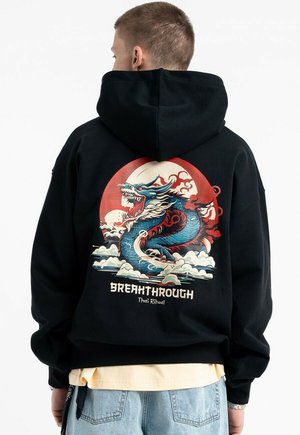 Osoba w czarnej bluzie z kapturem, z kolorową, ryczącą niebieską grafiką smoka i czerwonym słońcem, oraz napisem "BREAKTHROUGH Thai Ritual" z tyłu.
