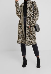 Długa kardiganka w panterkę z miękkiego materiału z dwoma przednimi kieszeniami, noszona na czarnym golfie i zestawiona z czarnymi szpilkami za kostkę.