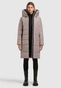 Zateplený dlhý kabát svetlo šedej farby s vysokým golierom a nadmerne veľkou kapucňou, s predným zipsom a bočnými vreckami. Nosený cez čierny outfit.