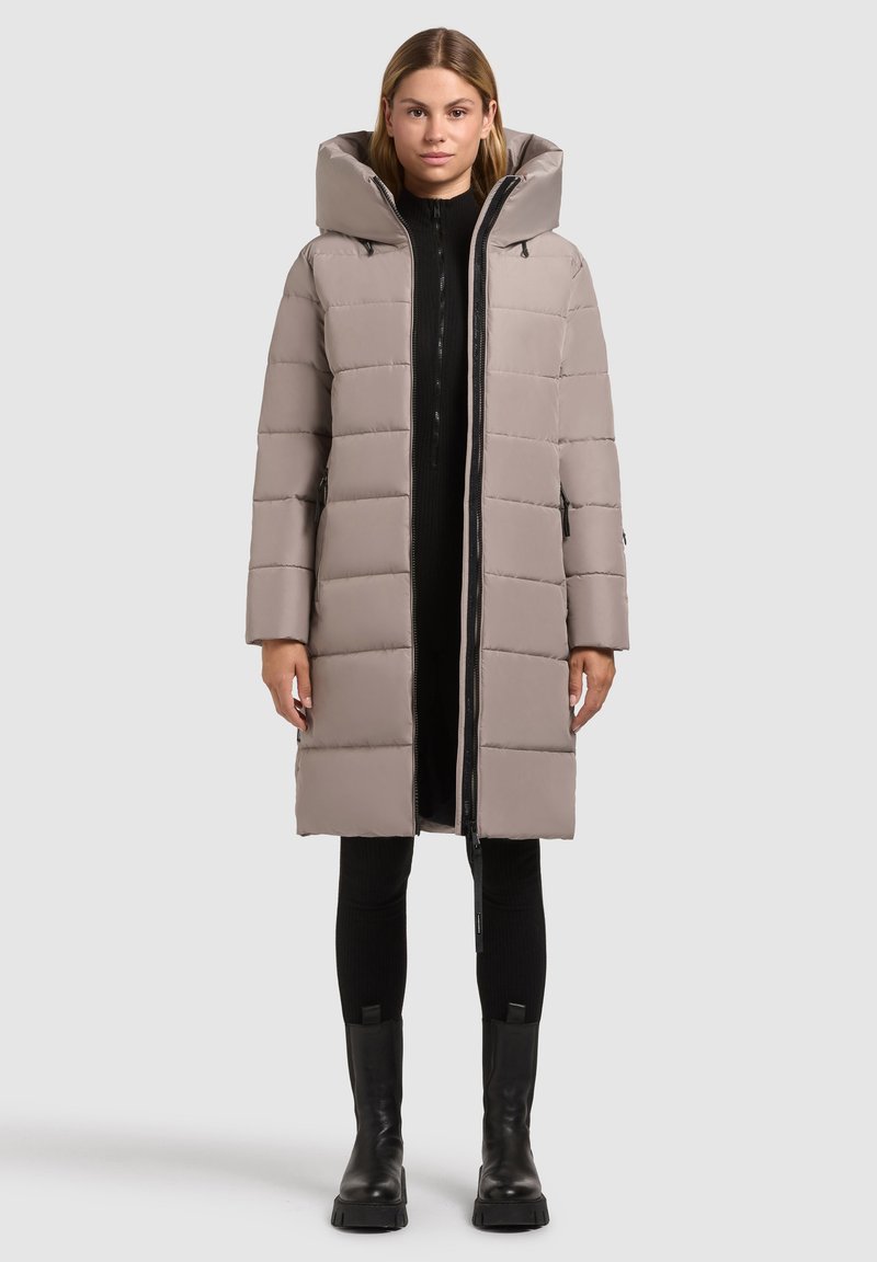 Zateplený dlhý kabát svetlo šedej farby s vysokým golierom a nadmerne veľkou kapucňou, s predným zipsom a bočnými vreckami. Nosený cez čierny outfit.