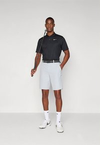 Polo preta com colarinho, calções cinza claro, cinto preto e sapatilhas brancas. Taco de golfe na mão direita. Fundo simples.