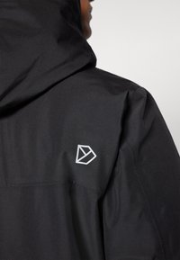 Černá kapucňová bunda s matným povrchem, s bílým geometrickým logo na rameni. Hladká textura, minimalistický design.