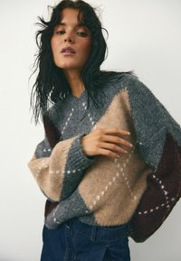 Paukščių megztinis su geometriniu raštu pilkos, smėlio ir bordo spalvų; pasižymi tekstūruota apdaila ir kontrastingomis spalvomis su baltomis detalėmis.