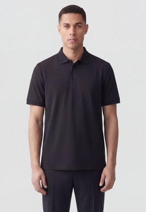 Uomo che indossa una polo nera semplice a maniche corte con un piccolo logo in rilievo, in piedi davanti a uno sfondo chiaro e uniforme.