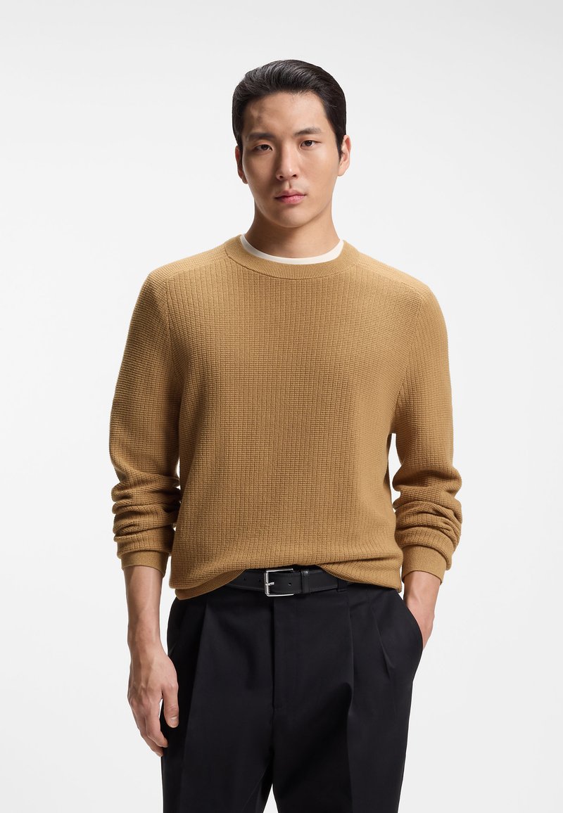 Jeune homme aux cheveux courts noirs portant un pull texturé couleur beige, un pantalon noir et une ceinture, debout avec une main dans la poche, sur fond blanc.