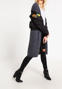 Svart bomberjacka med färgglada lappar, grå lång stickad cardigan, svarta leggings och ankelstövlar. Innehåller revor på leggings.