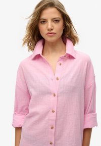 Camisa de botones rosa en un tejido ligero y texturizado. Presenta un collar clásico y botones de madera, con las mangas arremangadas hasta los codos.