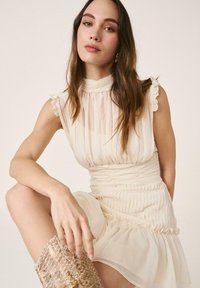 Femme aux longs cheveux bruns portant une robe crème sans manches et des bottes hauteur genoux en cuir de serpent, posant avec un genou levé sur un fond neutre.