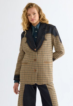 Jeune femme aux cheveux blonds ondulés porte un blazer pied-de-poule avec col en denim foncé et pantalon assorti sur fond blanc.