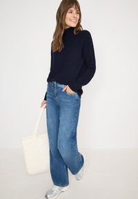 Pull marin, jean bleu à larges jambes et baskets en argent. Le mannequin tient un sac en toile matelassé, de couleur blanc cassé. Texture lisse, design décontracté.