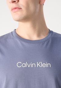 Gaiši zils kokvilnas T-krekls ar gludu tekstūru un īsām piedurknēm, ar "Calvin Klein" baltiem burtiem drukāts uz krūtīm.