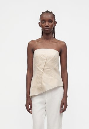 Femme aux cheveux tressés portant un haut beige sans bretelles avec un ourlet asymétrique et un pantalon blanc, debout devant un fond uni.