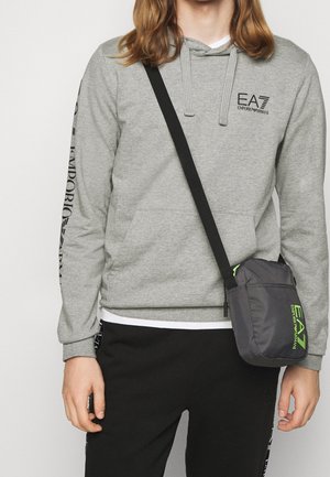 Grå sweatshirt med hætte, kengurulomme og sort EA7-logo, parret med sorte bukser og en lille sort skuldertaske med grøn detalje.