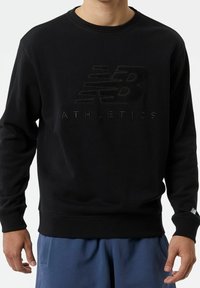 Felpa nera caratterizzata da un logo testurizzato e la scritta "ATHLETICS". Polsini e orlo a coste, tessuto morbido, vestibilità rilassata. Abbinata a pantaloni blu.