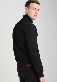 Giubbotto bomber nero con colletto alto, polsini ed elastico in vita. Tessuto liscio e opaco con spalle senza cuciture, indossato sopra jeans scuri.