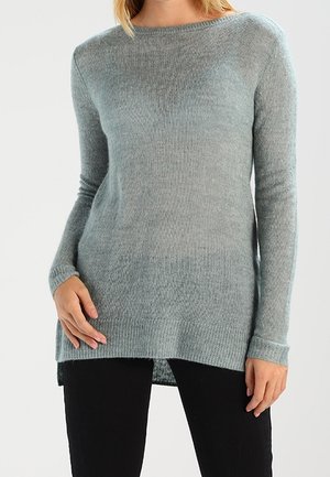 Frau trägt einen hellgrauen, langärmeligen Strickpullover mit rundem Ausschnitt und schwarze Hose vor neutralem Hintergrund.