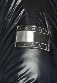 Tissu noir brillant avec un patch rectangulaire noir et argenté portant le texte "TOMMY JEANS" et des blocs de couleur texturés.