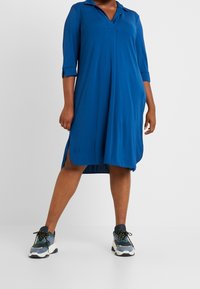 Woman wearing a blue knee-length dress with three-quarter sleeves and side slits, paired with blue and white chunky sneakers.