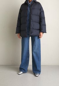Puffermantel in Dunkelblau, gesteppter Design mit großen Paneelen, kombiniert mit weiten blauen Jeans und silbernen spitzen Schuhen.