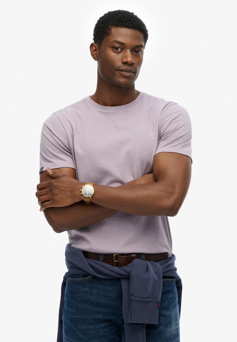 T-shirt en coton violet clair à manches courtes, col rond, avec une petite poche noire sur la manche. Montre en cuir marron et pull navy noué autour de la taille.