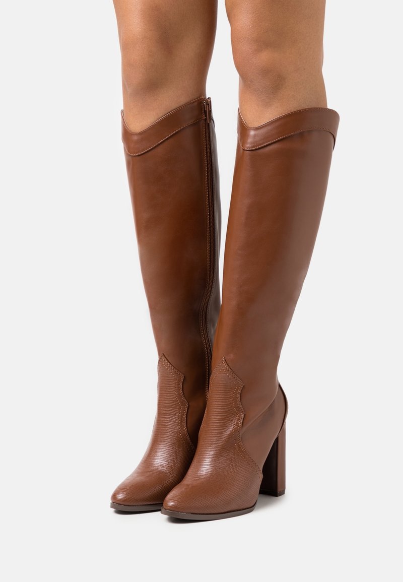 Bottes en cuir marron jusqu’au genou avec bout texturé et fermeture éclair latérale, portées sur jambes nues, avec des talons blocs hauts.