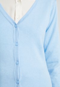 Světle modrý svetr s výstřihem do V vyrobený z měkké pleteniny, s předním zapínáním na knoflíky a hladkou texturou.