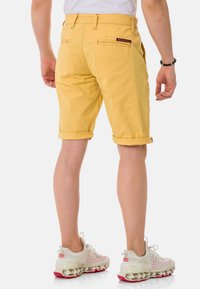 Gelbe Baumwollshorts mit aufgerollten Säumen, ausgestattet mit einer Gesäßtasche und roten Akzenten. Getragen mit hellfarbigen Sportschuhen.