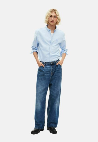 Persona rubia vestida con una camisa azul claro abotonada y mangas arremangadas, jeans anchos azules y zapatos negros, de pie con las manos en los bolsillos.