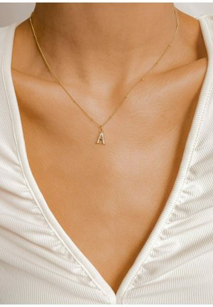 Goldene Halskette mit einem mit Kristallen besetzten Buchstaben "A", zarte Kette mit kleinen runden Akzenten, getragen über einem strukturierten, cremefarbenen Oberteil.