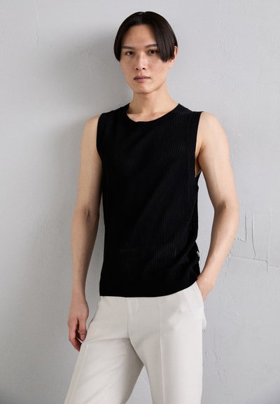 Black sleeveless ribbed top with a round neckline, worn with light-colored pants, featuring a subtle texture and form-fitting design.