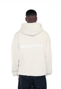 Béžová mikina s kapucňou s veľkým bielym logom na chrbte, vo voľnom strihu, vyrobená z mäkkej bavlny. V kombinácii s tmavými džínsami.