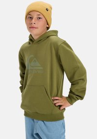 Vihreä huppari, jossa on etutasku ja logo; yhdistettynä vaaleansinisiin farkkuihin ja keltaiseen ribbimekko-pipoon.